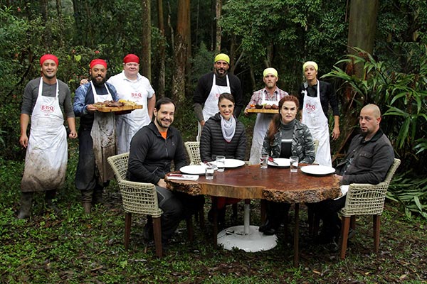 na foto, jurados aparecem sentados e os cozinheiros em pé segurando os pratos que assaram para a prova em grupo