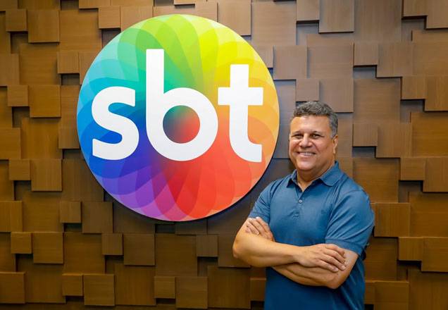 De braços cruzados, Téo José sorri para a foto com o logo do sbt ao fundo
