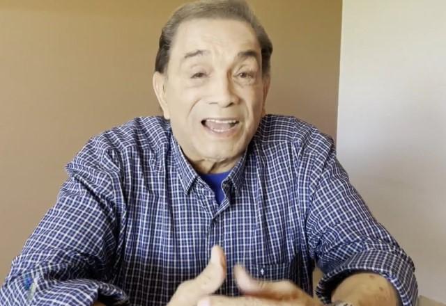 Dedé Santana sentado em mesa com fundo laranja