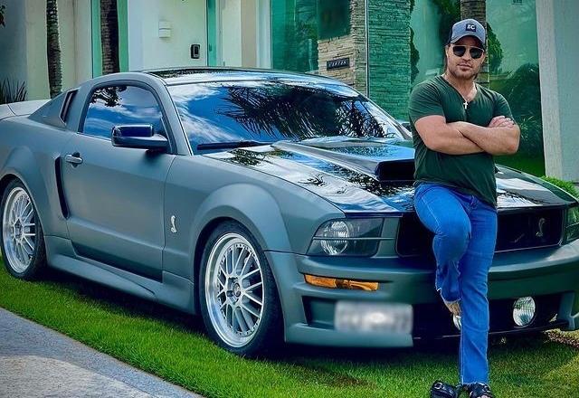 Eduardo costa vestido de boné, calça jeans, camisa verde e óculos escuros apoiado em seu carro, um mustang cinza