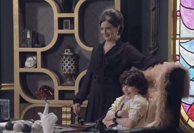 Norma, em pé, sorri ao lado de Felipe, que está sentado na cadeira da diretora, também sorrindo