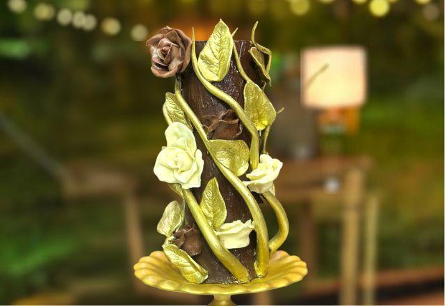 Na foto temos um bolo alto e sofisticado, decorado com rosas e folhas de chocolate modelado, detalhes dourados e acabamento inspirado em uma roseira.