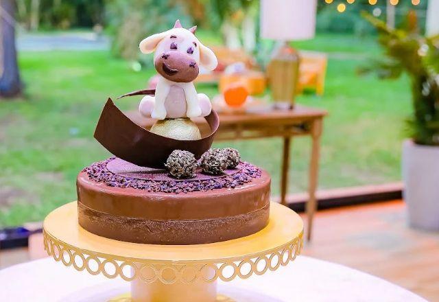 Na foto temos um entremet de chocolate com acabamento espelhado, decorado com uma escultura de vaca em chocolate e detalhes dourados.
