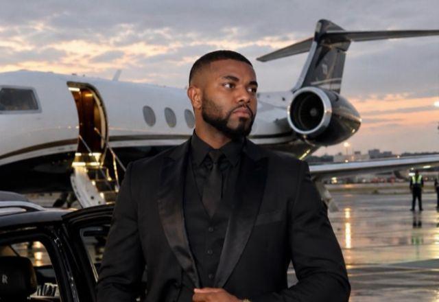 Davi Brito posa com uma aeronave ao fundo. Ele veste um blazer preto e a expressão facial é séria.