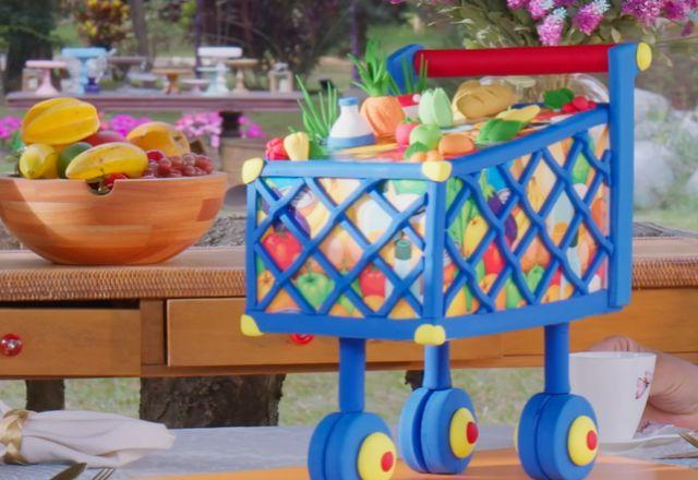 Na imagem temos um bolo em formato de carrinho de compras cheio de frutas e verduras. Toda a estrutura e produtos feitos com papel arroz e pasta americana.