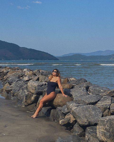 Giulia Costa inclinada em uma pedra na praia. Ela veste um maiô preto.