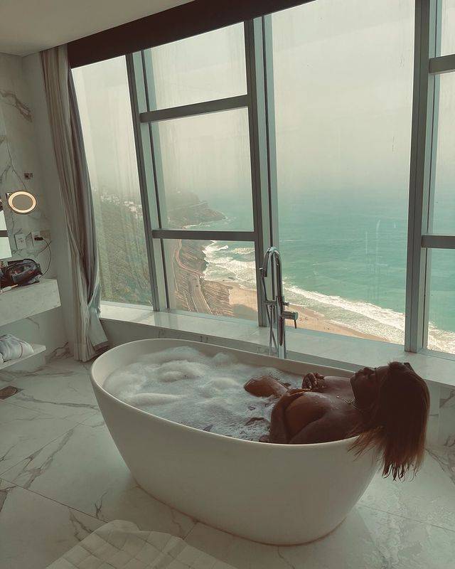 A cantora Jojo Todynho dentro de uma banheira de um hotel de luxo. Pela janela dá pra ver uma praia carioca.