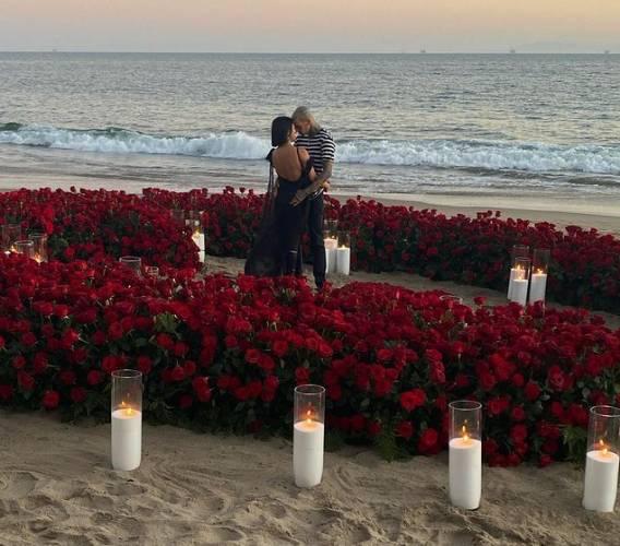 Kourtney de vestido preto, Travis de calça preta e camisa listrada. Os dois abraçados dentro de um coração gigante de flores com velas em uma praia. 