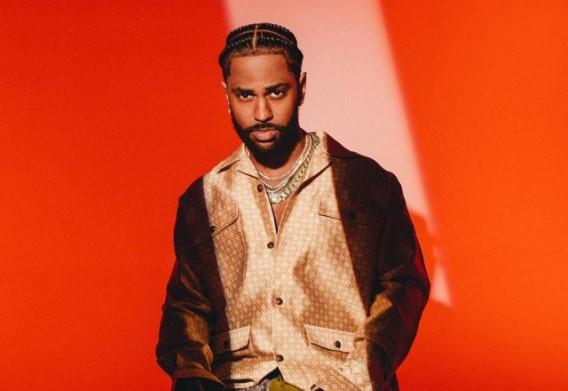 Rapper Big Sean usando uma camisa dourada e uma jaqueta marrom em um fundo vermelho