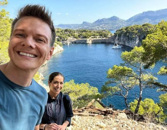 Michel Teló e Thais Fersoza no Sul da França em uma trilha com uma paisagem do mar e de árvores ao fundo