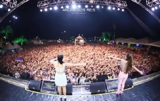 Maiara e Maraisa de costas, em cima do palco, catando com as mãos para o alto. De fundo a plateia repete o gesto das cantoras.