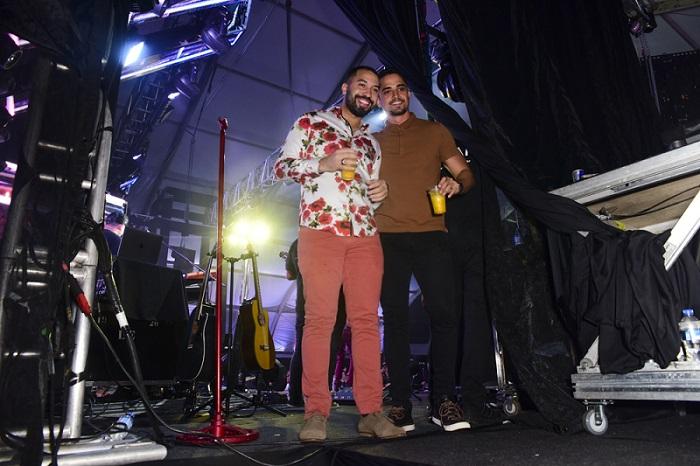 Gil e Lucas sorriem para a foto e seguram copos de bebida. Ao fundo, bastidores do show.