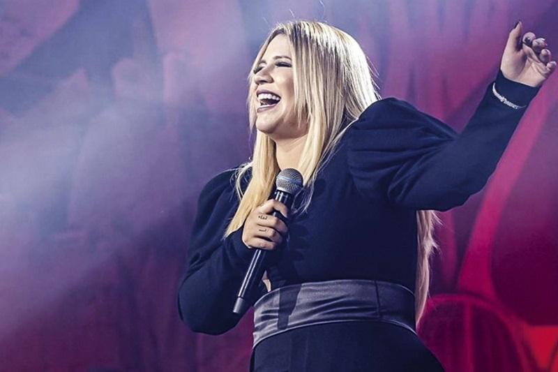 No palco, Marília Mendonça canta com uma das mãos para cima, enquanto segura o microfone com a outra.