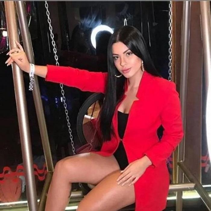 Rayane Figliuzzi posa para foto sentada em um balanço. Ela usa roupas vermelhas e pretas.