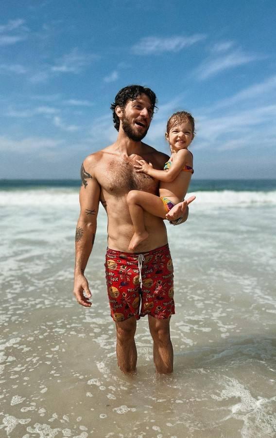 Chay Suede aparece só de shorts de praia, na beira do mar e segurando sua filha Maria no colo, os dois estão sorrindo