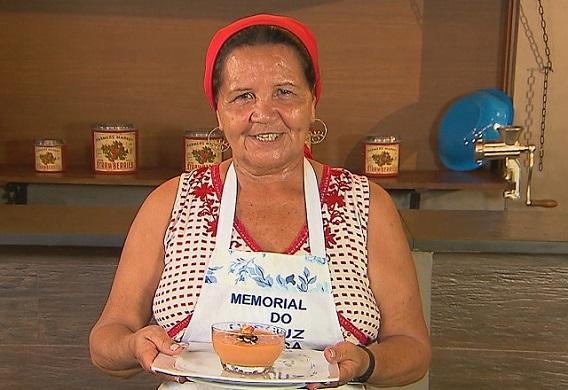 Dona Lia posa com uma faixa vermelha no cabelo e um avental escrito Memorial do Cuscuz. Ela segura uma bandeja com um doce de goiaba servido em um recipiente de vidro transparente