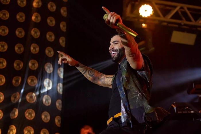 Gusttavo Lima sorri com as mãos para o alto, em cima do palco, durante um show.