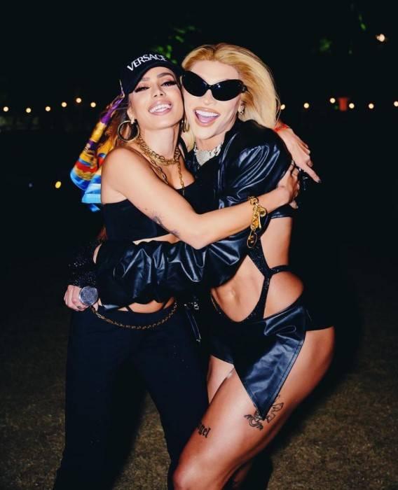 Anitta e Pabllo Vittar abraçadas e sorrindo, ambas estão com roupas em tons de preto, Anitta com um lenço no cabelo e Pabllo com um óculos escuros