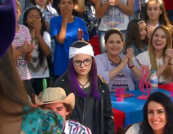 Isabela olhando com raiva para o palco, atrás tem pessoas sorrindo e em primeiro plano aparece um pouco de Manuela de costas