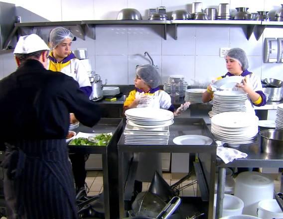 Joaquim, Felipe e Julia lavando pratos em uma cozinha de restaurante, em primeiro plano, aparece um garçom de roupa preta e de costas