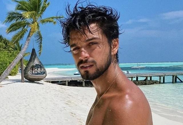 Na praia, Rodrigo Simas olha para foto fazendo carão