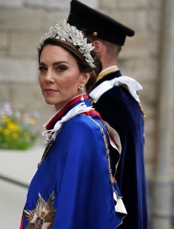 kate middleton usando uma tiara prata, brincos de pérolas, cabelo preso em coque e com uma capa azul com detalhes em vermelho. ela está de perfil