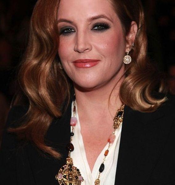 Lisa Presley posa sorrindo, usando uma camisa branca, blazer preto e colar