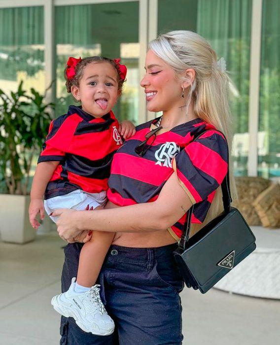 Karoline segura a filha no colo. Elas vestem a camisa do Flamengo