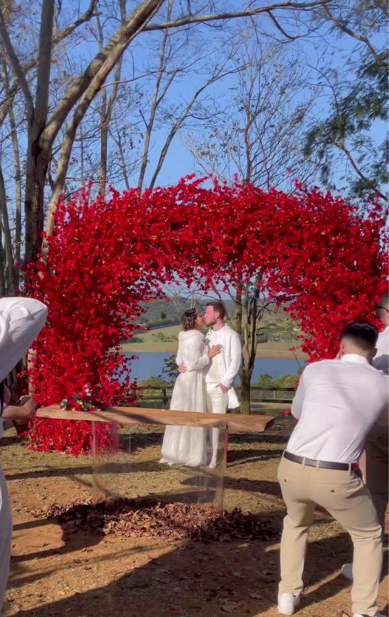 Maíra Cardi e Thiago Nigro embaixo de um arco de flores vermelhas, vestidos de branco e sendo fotografados